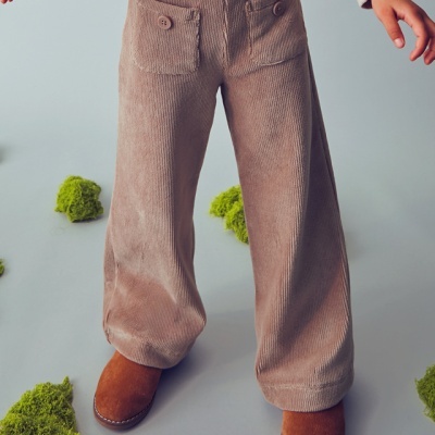 Criança a usar calças largas de veludo castanho claro e botas castanhas sobre fundo cinzento com musgo verde