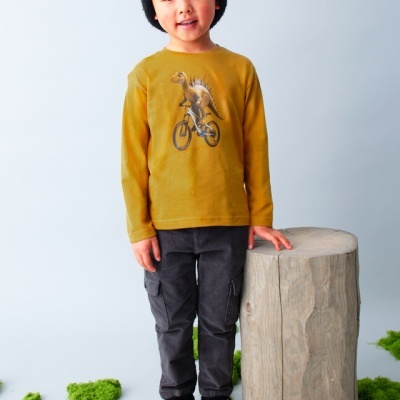 Menino com camisola amarela estampada, calças pretas, sapatos pretos e gorro preto, posando junto a um tronco de madeira e musgo verde.