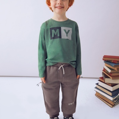 Menino ruivo com camisola verde e calças cinzentas, calçado desportivo, em fundo branco perto de pilha de livros.