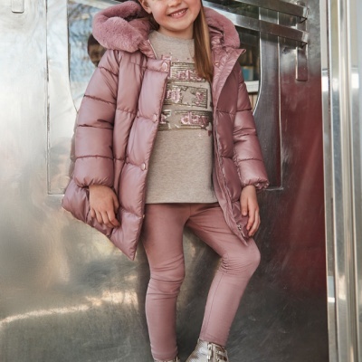 Menina com casaco acolchoado rosa, camisola bege, calças rosa e ténis prateados junto a porta metálica