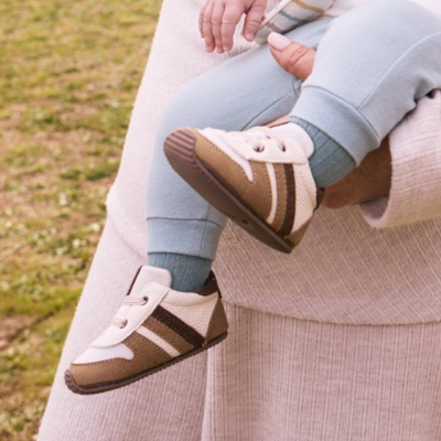 Sapatilhas para bebé branco e castanho com riscas castanhas, criança com calças azuis e meias verdes segura por adulto