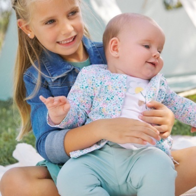 Criança com jaqueta de ganga segura bebé com casaco floral sentado ao ar livre
