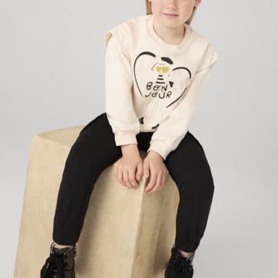 Menina com sweater bege e calças pretas sentada num bloco bege