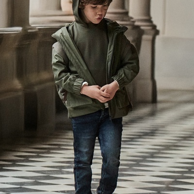 Menino vestido com camisola e casaco verdes, jeans azuis e sapatilhas pretas, num corredor com colunas e chão xadrez.