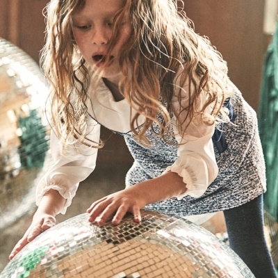Criança loira com vestido azul e branco a tocar numa grande bola disco prateada