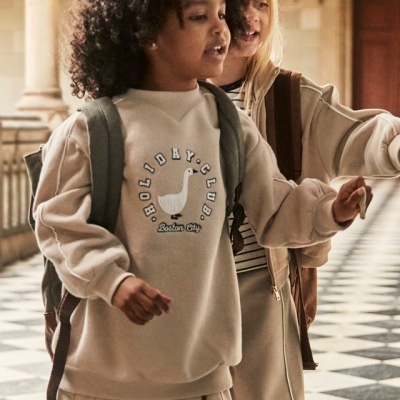 Duas crianças com roupas casuais bege e mochilas, num espaço com colunas e pavimento xadrez.