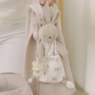 Boneco de peluche coelho bege branco com chupeta pendurado em tecido bege.