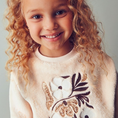 Camisola creme felpuda com bordado floral usada por criança sorridente.