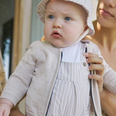 Bebé de chapéu riscado branco e bege, vestido branco riscado, cardigan bege, segurado por mulher ao ar livre.