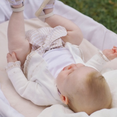 Bebé vestido com casaco branco, calções às riscas bege e brancas e sapatos beges com detalhes lilás deitado numa camada branca com relva ao fundo