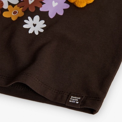 Detalhe de tecido castanho com estampado floral e etiqueta preta com texto branco.