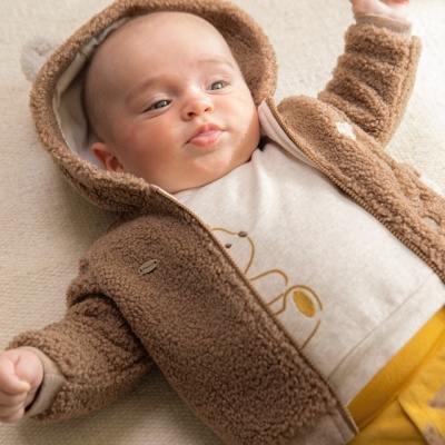 Bebé com casaco castanho felpudo, camisola creme com desenho e calças amarelas deitado sobre superfície clara