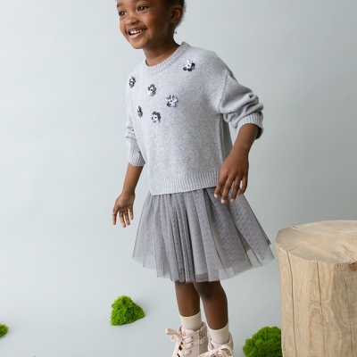 Criança com vestido cinzento e botas cor de rosa em fundo neutro com musgo e tronco de madeira.