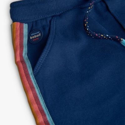 Detalhe de calça azul com faixa lateral colorida e botão com texto