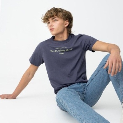 Jovem sentado com camiseta azul escuro com texto, jeans azul claro e ténis brancos