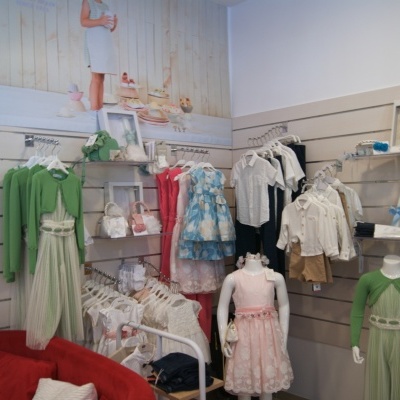 Loja de roupa infantil com vestidos coloridos em cabides e manequins, parede branca com painéis e figura de criança em miniatura.