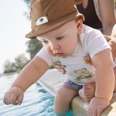 Bebé com chapéu de urso e t-shirt com ursinho sentado à beira de piscina