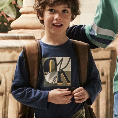 Camisola azul escuro criança com estampado CK, mochila castanha, camisa às riscas amarrada à cintura, colunas de pedra e plantas.