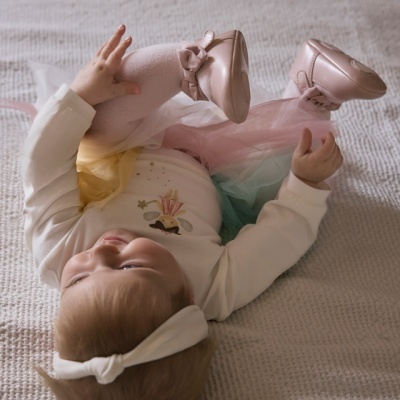 Bebé deitada com vestido de tule colorido, sapatos rosa e faixa branca na cabeça sobre manta bege.