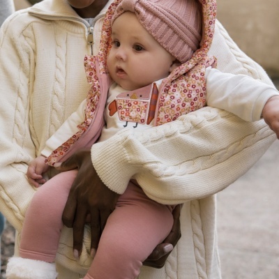 Bebé com roupa rosa e colete floral, segurado por adulto com camisola creme de malha