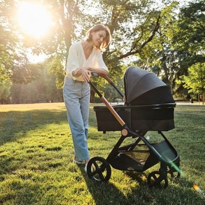 Mulher empurrando carrinho de bebé preto num parque gramado com árvores e luz solar