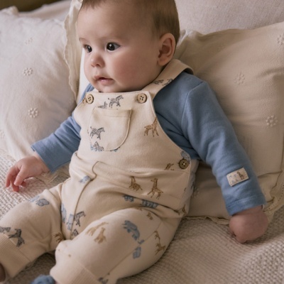 Bebé com macacão creme estampado e camisa azul sentado em cama creme