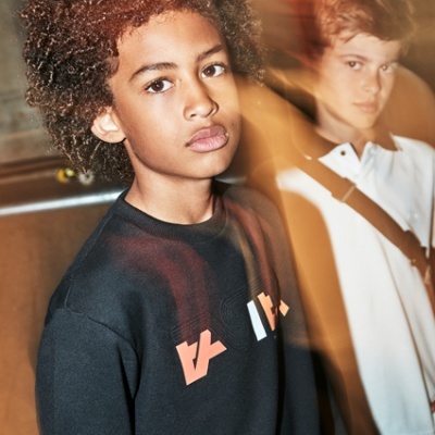 Dois rapazes adolescentes em roupa casual, um de preto com gráficos, outro de branco com alça castanha.