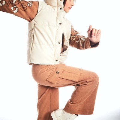 Criança vestida com colete branco, calças castanhas e botas brancas, e um gorro castanho, em pose dinâmica.