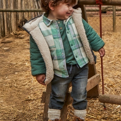 Menino com casaco verde, camisa xadrez, jeans e botas em exterior com chão de palha