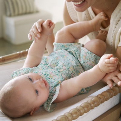 Bebé deitado em cama de madeira com roupa floral e mulher sorridente a segurá-lo pelas mãos