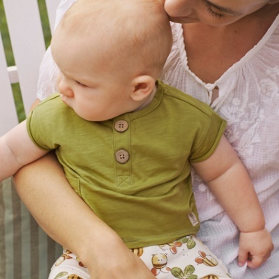 Bebé vestido com roupa verde e calções estampados no colo de mulher com blusa branca sentados em cadeira ao ar livre