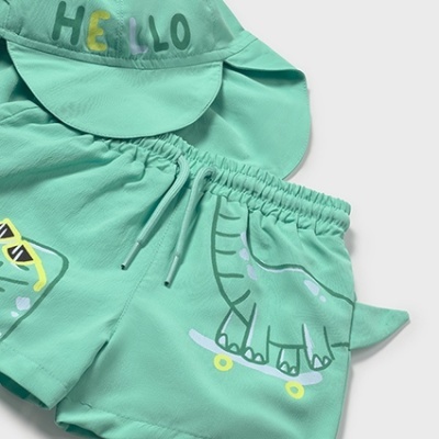 Conjunto de roupa infantil verde com bermuda e boné estampados com dinossauros e a palavra HELLO.