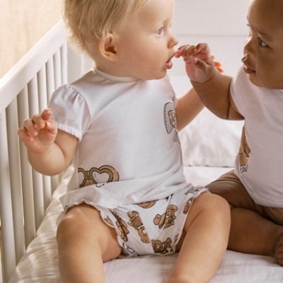 Dois bebés vestidos com roupa branca com padrões castanhos sentados numa cama branca