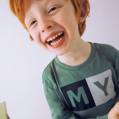 Camisola verde de manga comprida com a palavra MY em blocos coloridos, criança ruiva sorridente.