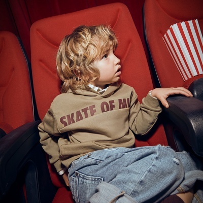 Criança sentada numa cadeira vermelha de cinema com camisola bege e jeans azuis ao lado de balde de pipocas vermelho e branco
