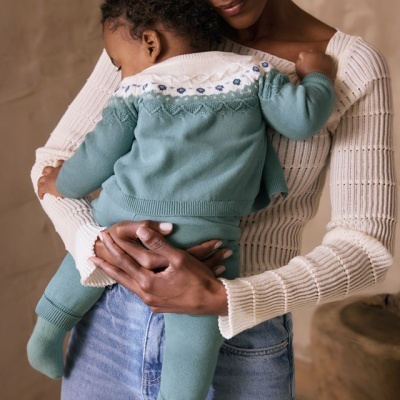 Bebé vestido com conjunto azul-esverdeado com padrão, segurado por mulher com camisola branca e jeans