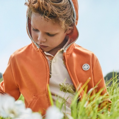 Menino de casaco laranja com capuz e polo bege com padrão floral ao ar livre entre relva e flores brancas