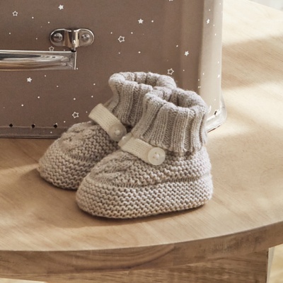 Botinhas de bebé creme tricotadas em mesa de madeira com mala cinzenta de estrelas ao fundo