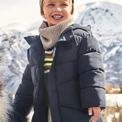 Criança com casaco azul, camisola riscada, gola e gorro bege, em paisagem nevada
