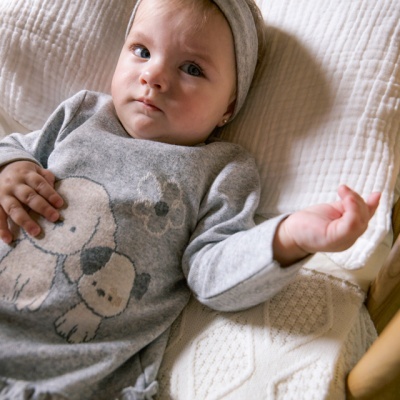 Bebé com vestido cinzento com desenho de cães e faixa de cabelo cinzenta deitada numa cama