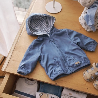 Casaco azul com capuz para criança sobre mesa de madeira com roupa dobrada na gaveta