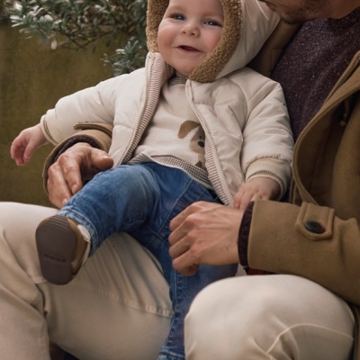 Bebé com casaco acolchoado bege e capuz de pêlo castanho sentado no colo de adulto com casaco castanho
