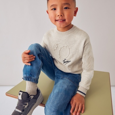 Criança vestindo camisola bege de malha com bordado, jeans azul e ténis cinzentos, sentada numa mesa escolar verde clara