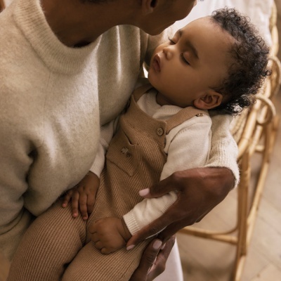 Bebé adormecido de macacão castanho claro e camisola branca segurado por adulto com camisola bege em cadeira de bambu