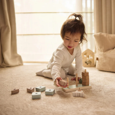 Criança a brincar com brinquedo de empilhar em madeira sobre carpete bege em sala com cortinas.