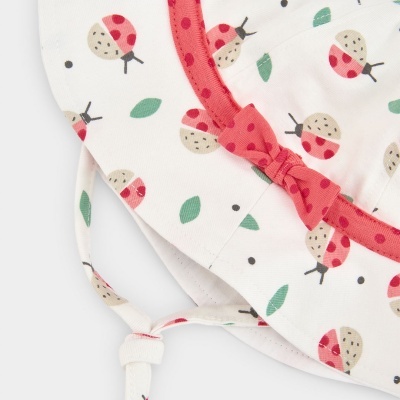 Chapéu de bebé branco com padrão de joaninhas vermelho e bege, folhas verdes, fita vermelha com bolinhas e laço.