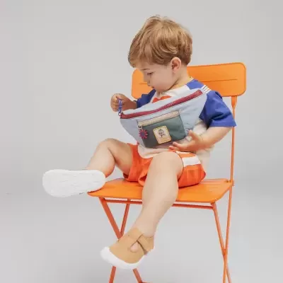 Criança com bolsa azul claro e verde sentada numa cadeira laranja.