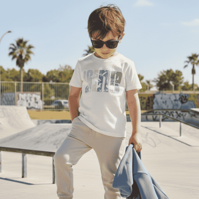 Menino de óculos de sol com t-shirt branca e calças bege num parque de skate