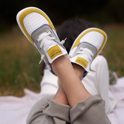 Sapatilhas de criança brancas com detalhes amarelos e cinzentos vistas em pernas cruzadas sobre manta ao ar livre