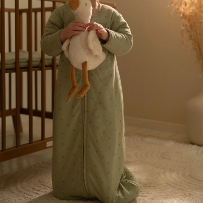 Criança com saco de dormir verde segurando pato de peluche em quarto com berço